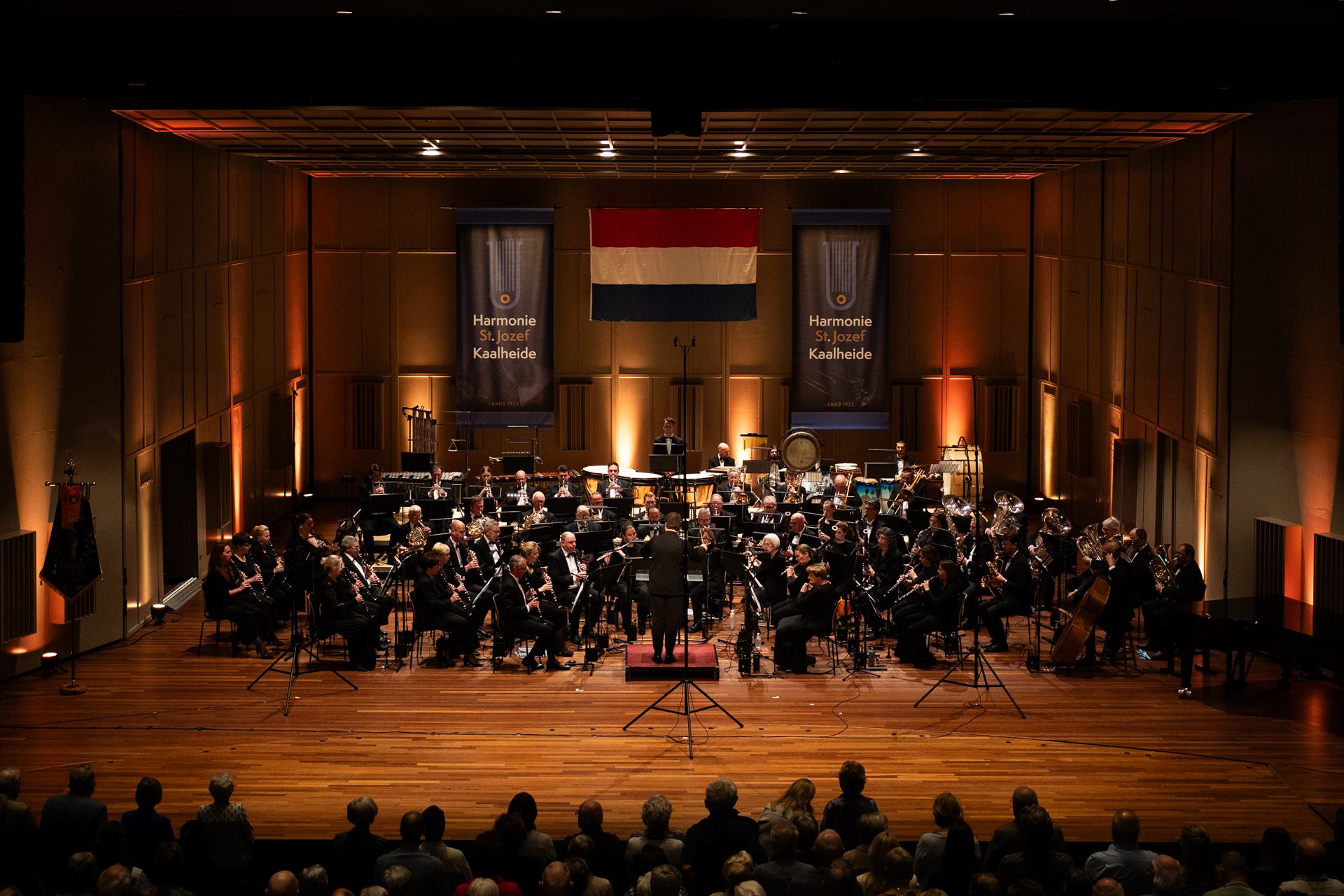 Dirigent Lars Corijn op de bok voor het voltallige orkest, met de vlag en de banners van de harmonie boven het podium
